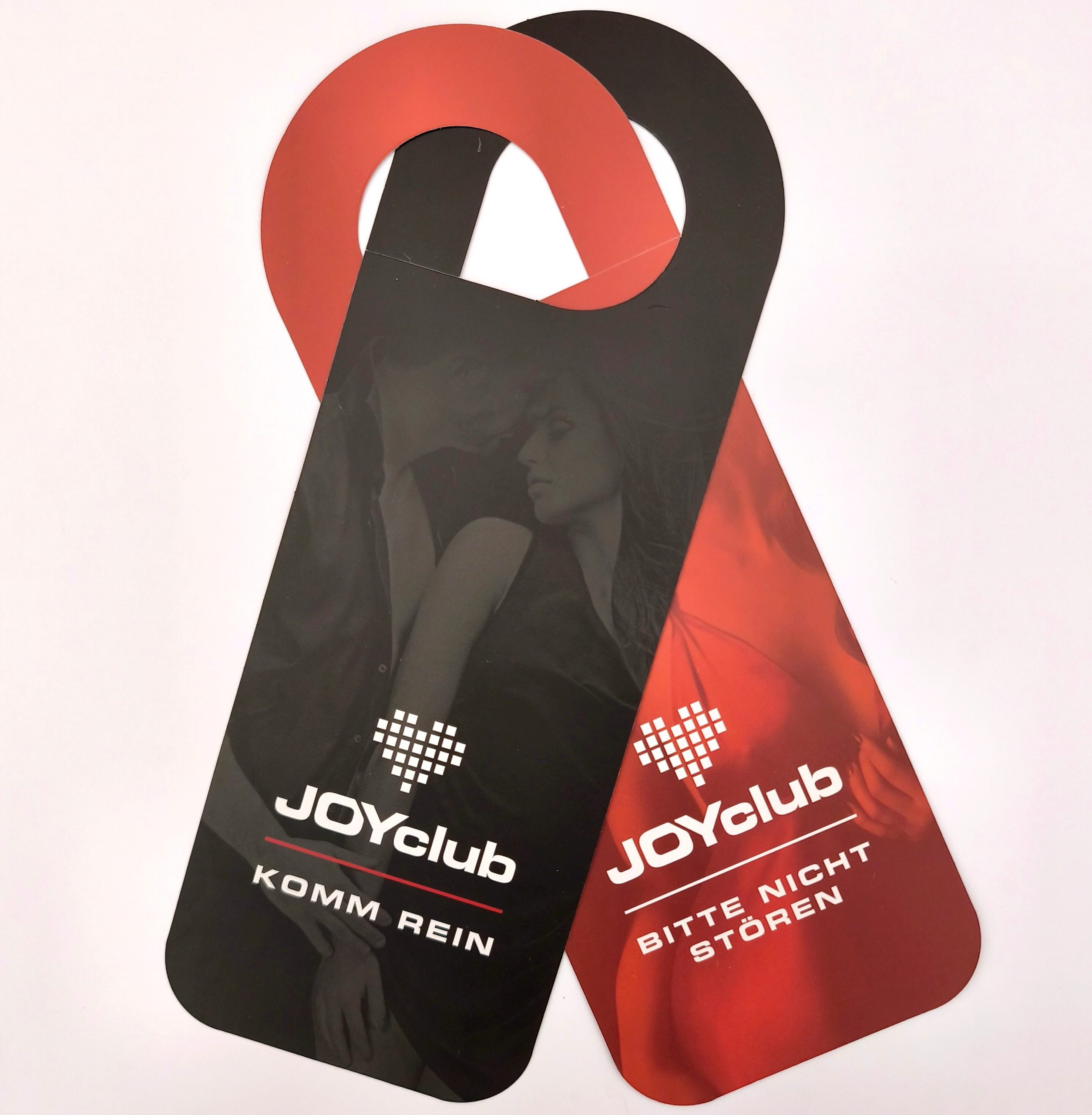 Zwei JOYclub Türhänger: einer schwarz mit „KOMM REIN“, einer rot mit „BITTE NICHT STÖREN“ und Paar-Motiv.