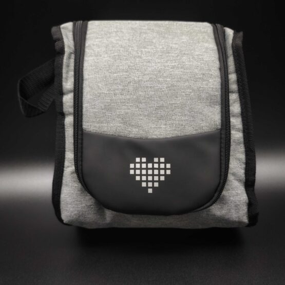 Eine stehende, graumelierte Kosmetiktasche mit schwarzem Reißverschluss und Tragegriff. Auf der Vorderseite befindet sich eine aufgesetzte, halbrunde, schwarze Tasche mit einem weißen, gepunkteten Herzsymbol in der Mitte. Der Hintergrund ist dunkelgrau bis schwarz.